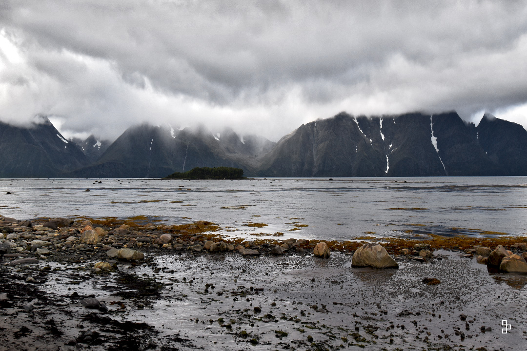 Un horizon montagneux , une petite ile dans ce fjord et en avant plan des petites roches. Le temps est gris et menaçant.