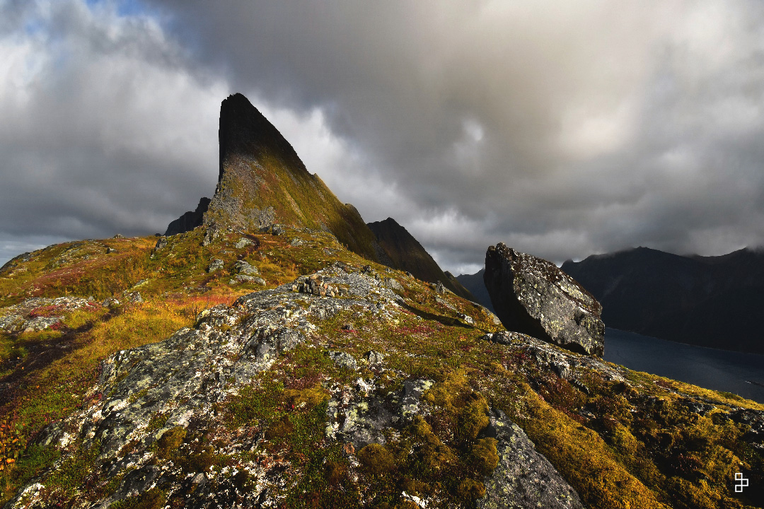 Un horizon montagneux , une petite ile dans ce fjord et en avant plan des petites roches. Le temps est gris et menaçant.