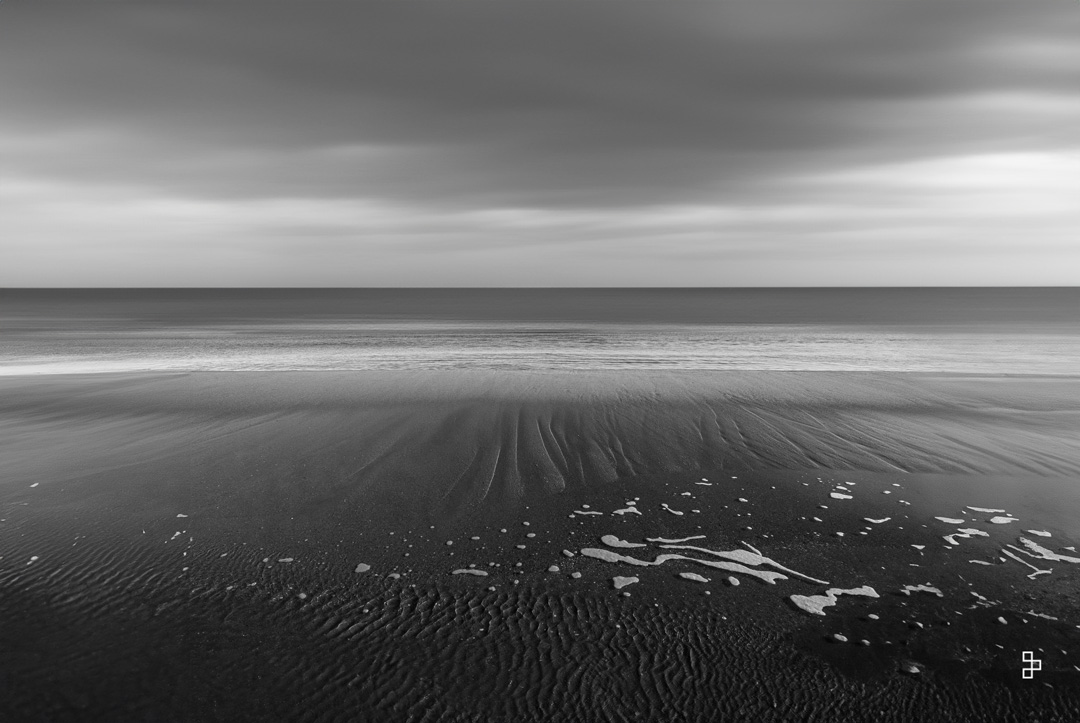 Un horizon montagneux , une petite ile dans ce fjord et en avant plan des petites roches. Le temps est gris et menaçant.